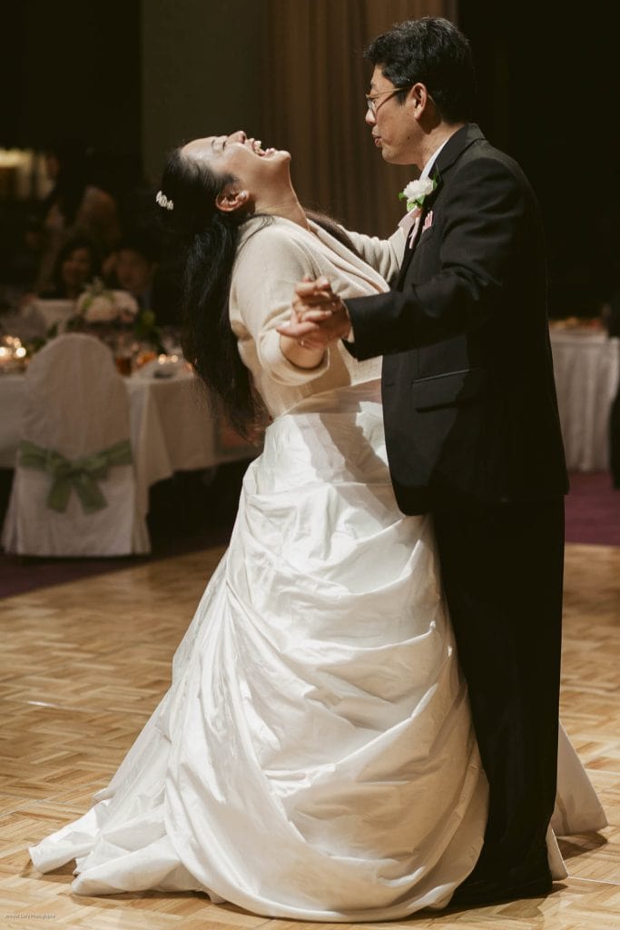 A bride in a white gown dances with an older man in a suit at a wedding reception, laughing joyfully. Guests sit at decorated tables in the softly lit background.