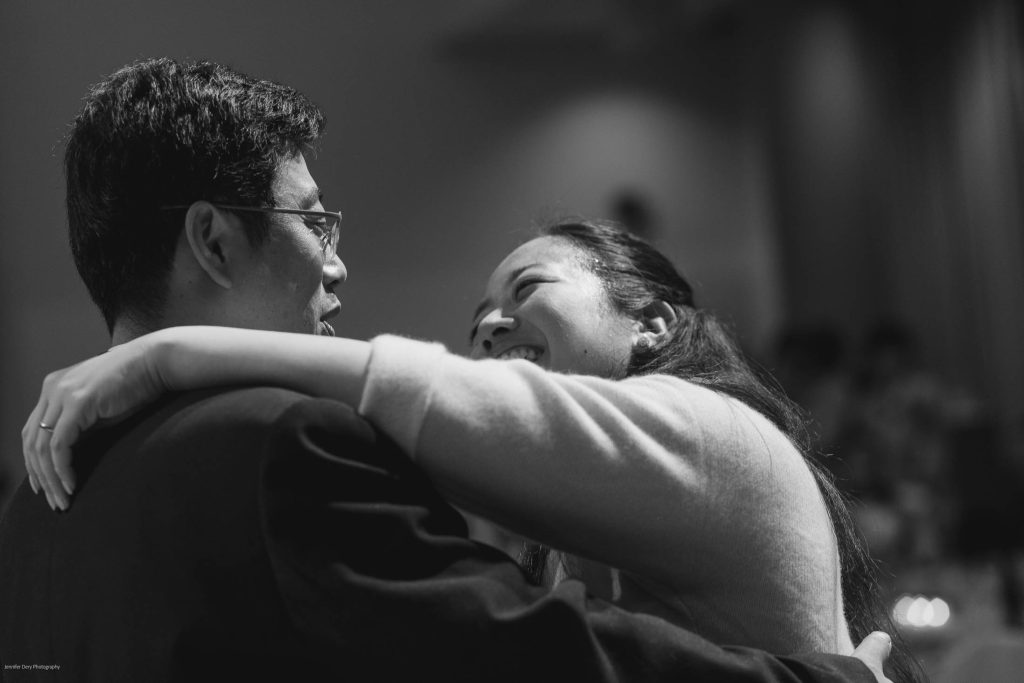 A black-and-white photo of a woman smiling joyfully with her arms around a man, both looking at each other closely, suggesting a moment of happiness and connection.