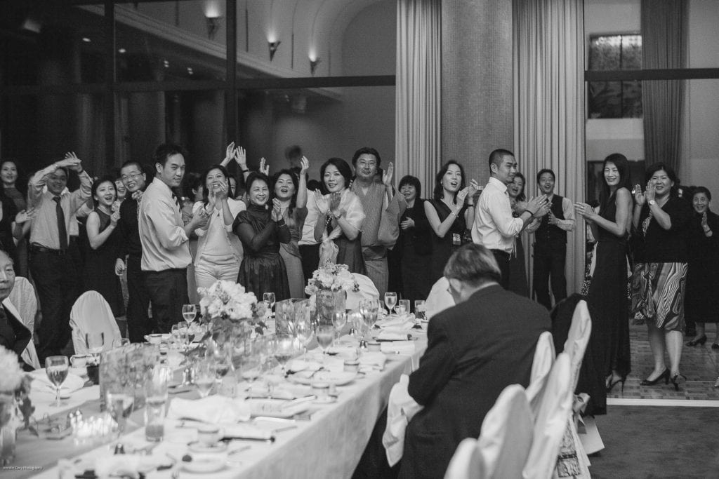 A group of people at a formal event, dressed in elegant attire, stand and cheer near a long, decorated dining table while others are seated, all in a well-lit room with large windows.