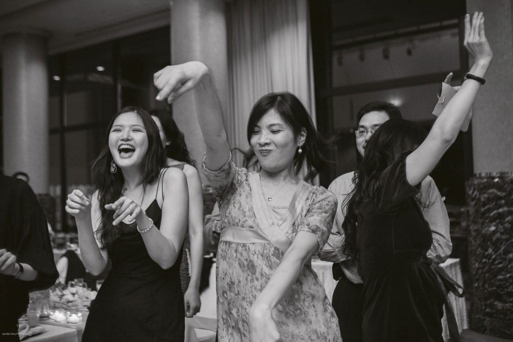 Four people are enjoying themselves at an indoor party. Three women are dancing energetically, smiling and laughing, while a man in glasses stands in the background, partially obscured. The photo is in black and white.