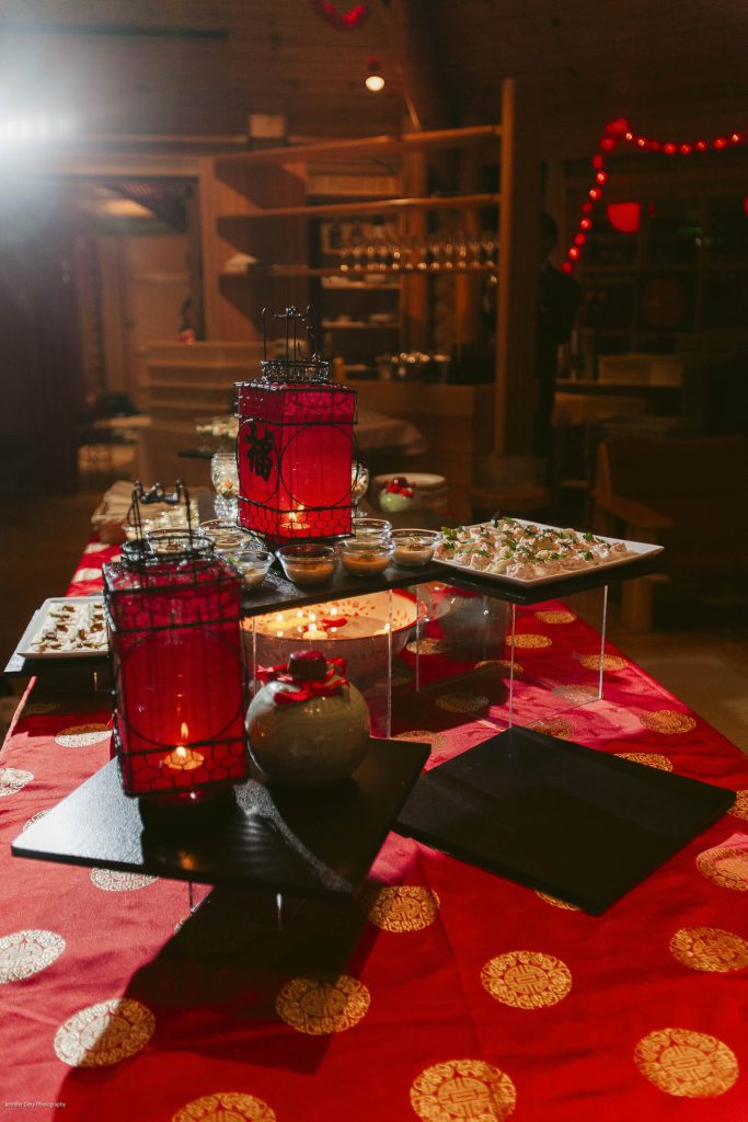 A red tablecloth with gold patterns is set with Asian-themed lanterns, candles, and a variety of plated appetizers, creating a festive atmosphere in a warmly lit room.