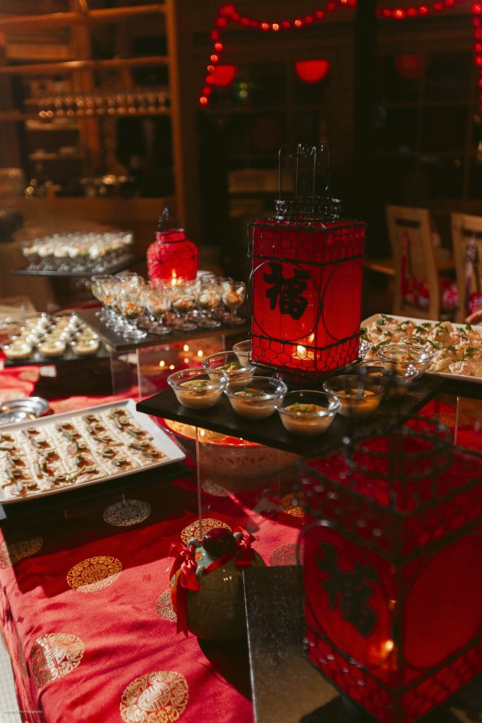A festive table decorated with red lanterns and gold-patterned cloth displays various small dishes and appetizers, set for a celebration in a warmly lit room.