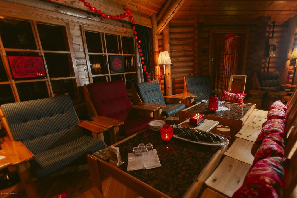 Cozy wooden cabin interior with armchairs, a long sofa, and tables set for tea. Warm lamps and red lantern decorations create a welcoming atmosphere. Large windows reveal evening or nighttime outside.