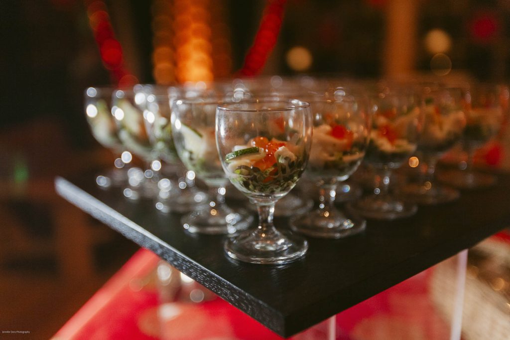 A close-up of several glass goblets filled with a colorful vegetable salad, arranged neatly on a black serving tray at a buffet or event. Warm lighting creates a festive atmosphere in the background.