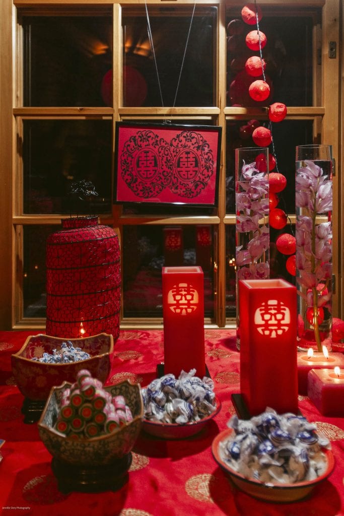 A festive table decorated with red candles, bowls of wrapped candies, floral arrangements, and red lanterns, set in front of a window with a double happiness symbol, evoking a traditional Chinese celebration.