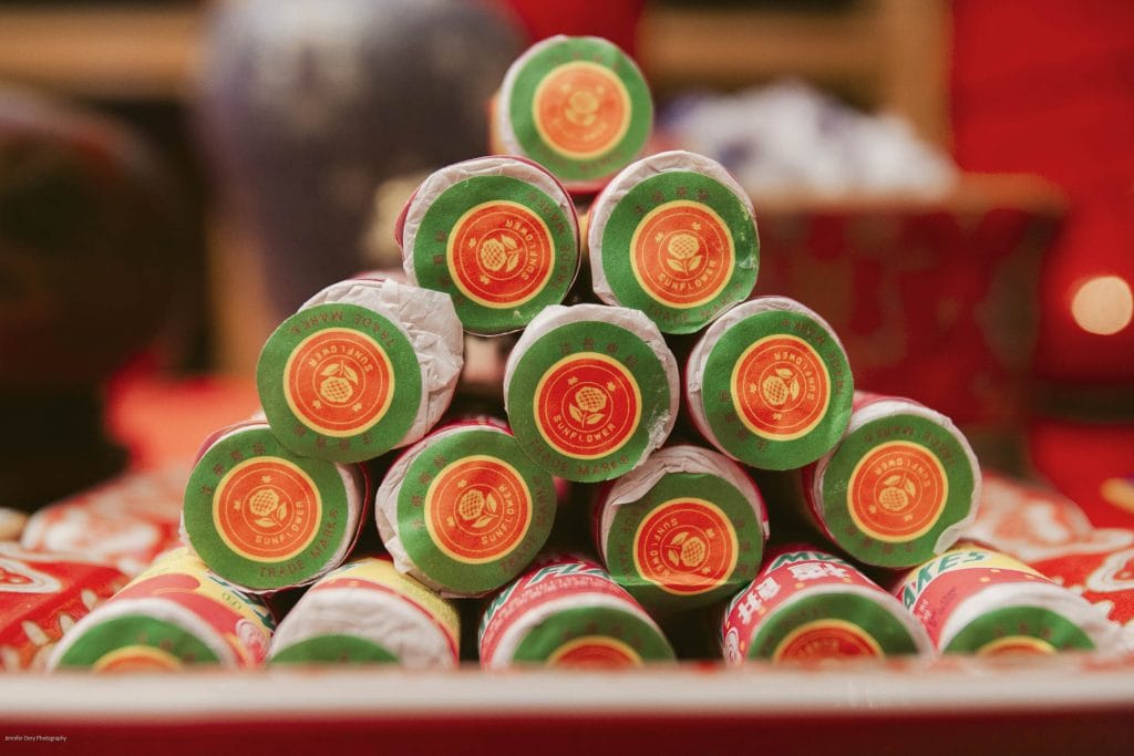 A close-up of a pyramid stack of rolled firecrackers wrapped in red and green paper, commonly used for Lunar New Year celebrations, set against a festive red background.