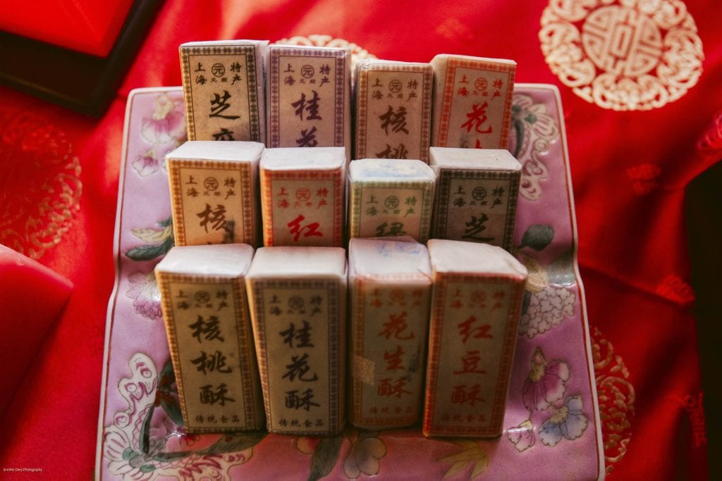 A plate of traditional Chinese pastries wrapped in colorful paper packaging sits on a red decorative cloth with intricate floral and Chinese character patterns, often used for festive occasions.