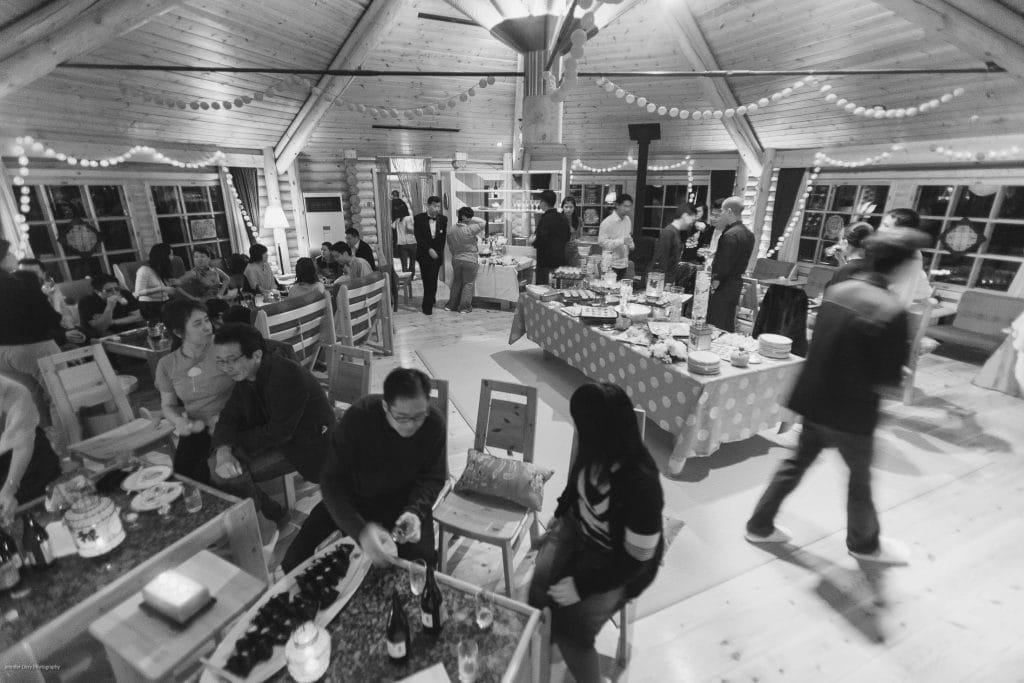 A black-and-white photo of people socializing and dining in a wooden, cabin-like venue decorated with string lights. Tables are set with food and drinks, and guests are seated or standing around the room.