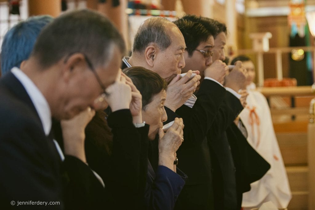 A group of people in formal attire stand in a line, holding small cups to their mouths, participating in a traditional Japanese ceremony inside a wooden building.