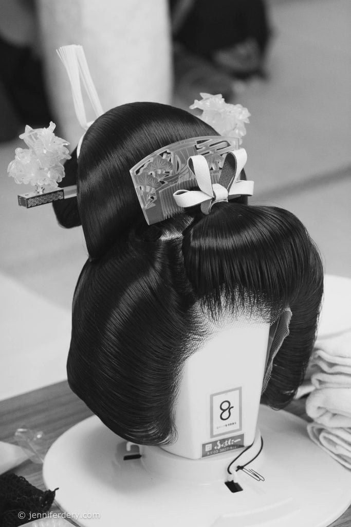 A traditional Japanese wig styled with ornate hairpieces, decorative combs, and floral accessories sits on a mannequin stand. The image is in black and white.
