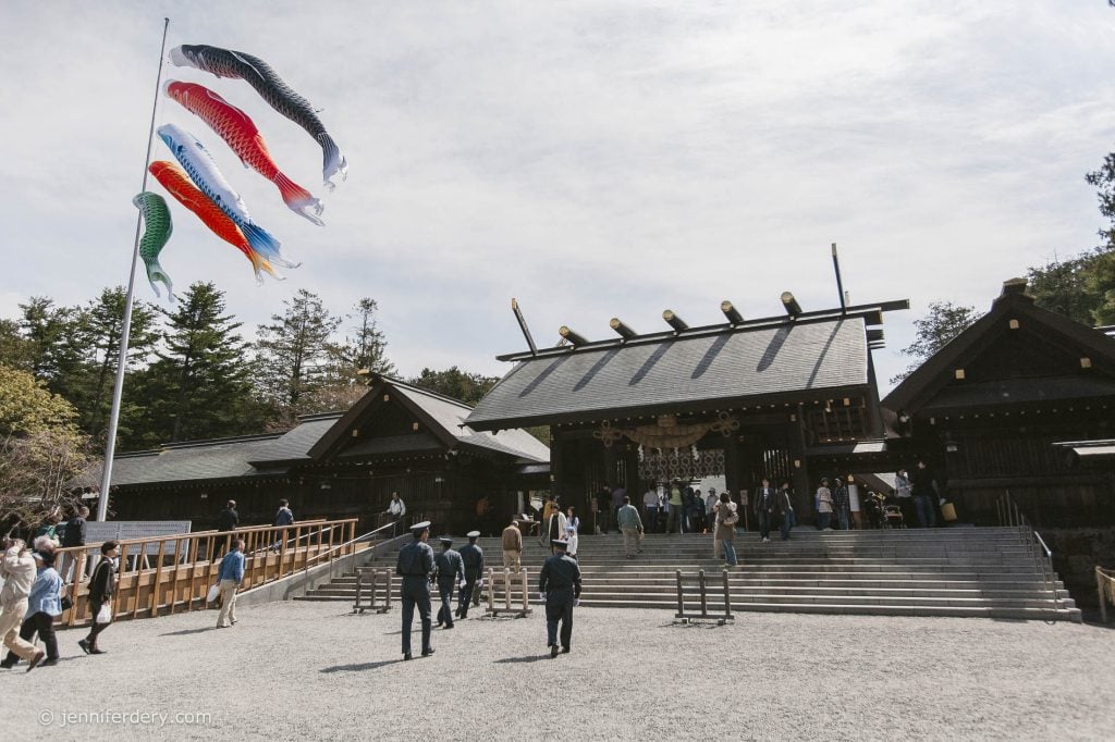 A group of people walk toward a traditional Japanese shrine. Colorful koi-shaped windsocks fly on a tall pole nearby, while trees and a blue sky are visible in the background.