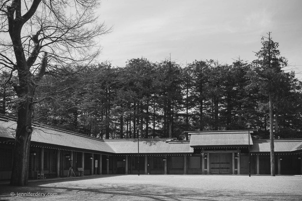 A black and white photo of a traditional Japanese courtyard surrounded by wooden buildings with tiled roofs, set against a backdrop of tall trees under a lightly clouded sky.