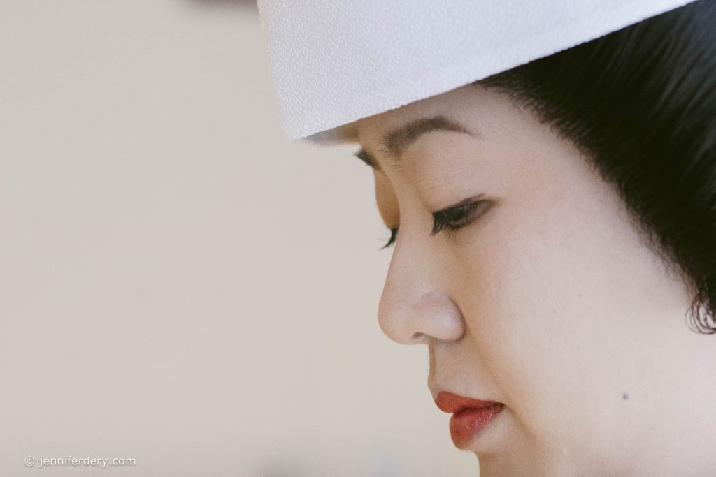 Close-up side profile of a woman with red lipstick and neatly styled black hair, wearing a white hat. The background is softly blurred, and the mood is calm and contemplative.