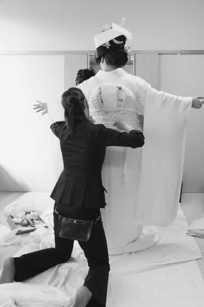 A woman in traditional Japanese bridal attire stands with her arms outstretched as another woman kneels and adjusts her kimono from behind in a bright, minimalist room.