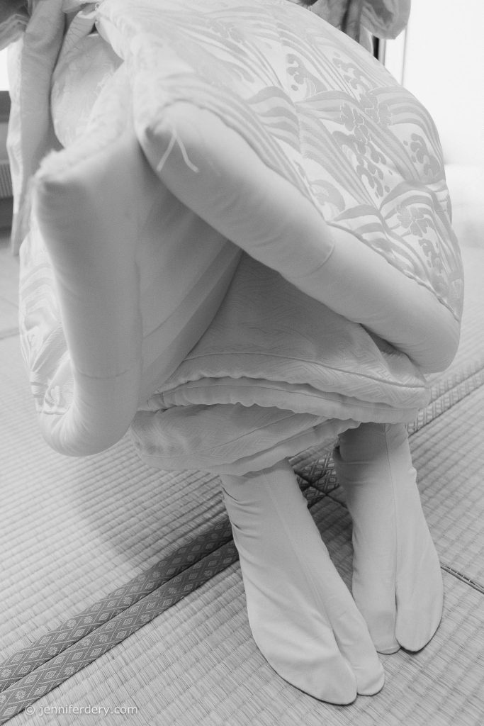 A black-and-white photo shows a person kneeling on tatami mats, wearing traditional Japanese bridal attire, including patterned fabric and white split-toe tabi socks.