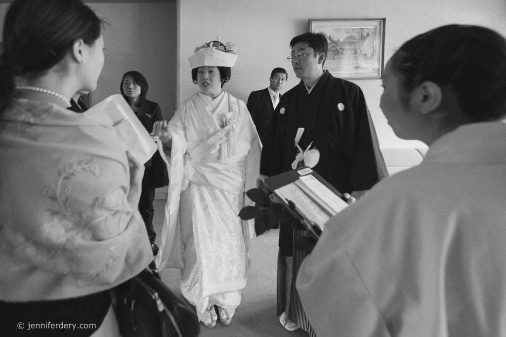 A bride in traditional Japanese wedding attire stands beside a groom in a formal kimono, surrounded by guests indoors, engaged in conversation. The scene appears candid and ceremonial.