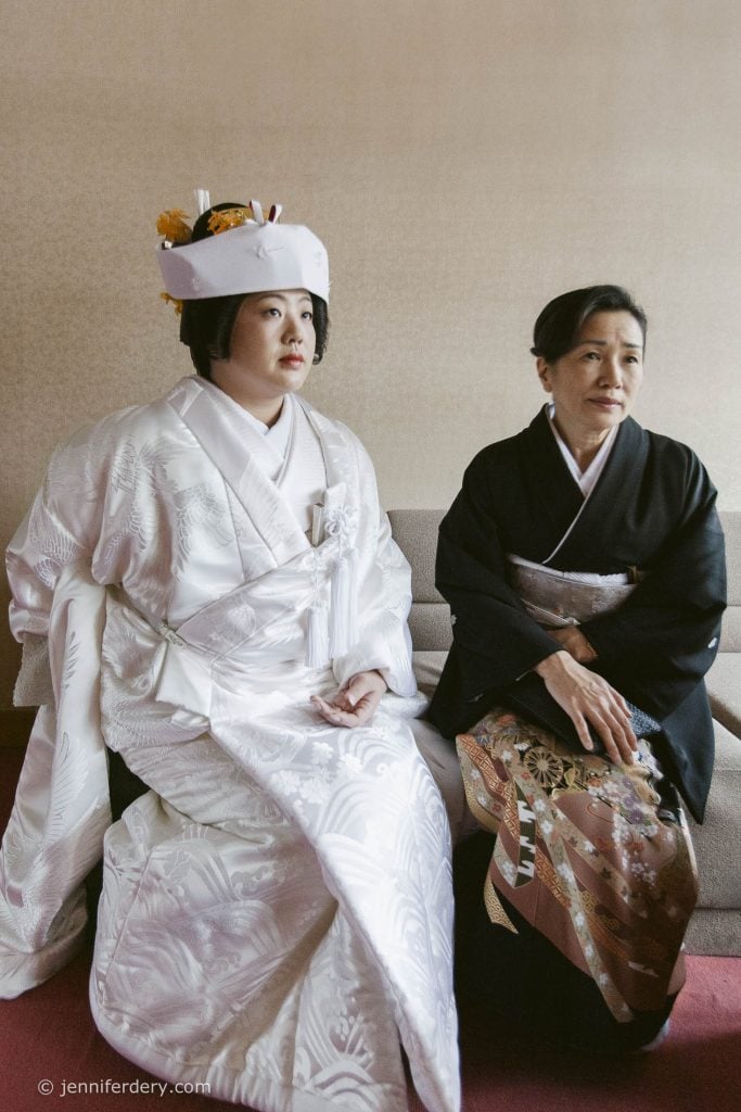A bride in a traditional white kimono and headdress sits beside an older woman in a black kimono with a patterned skirt, both looking to the side in a softly lit room.