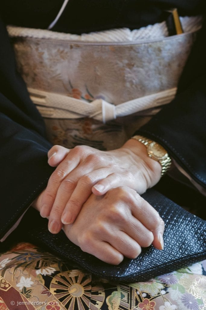 Close-up of a person’s hands resting on a black textured purse, wearing a gold wristwatch and a traditional kimono with floral patterns and a white obi belt.