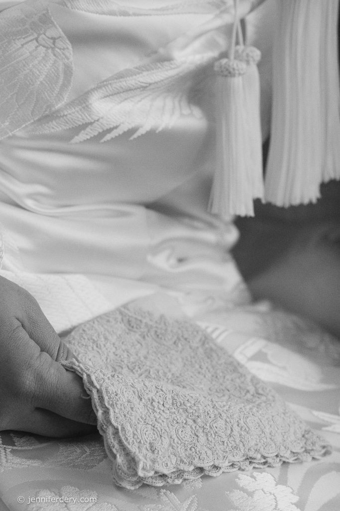 A close-up in black and white of a person wearing an embroidered garment with a textured lace accessory, possibly part of traditional attire, and holding fabric with delicate patterns.