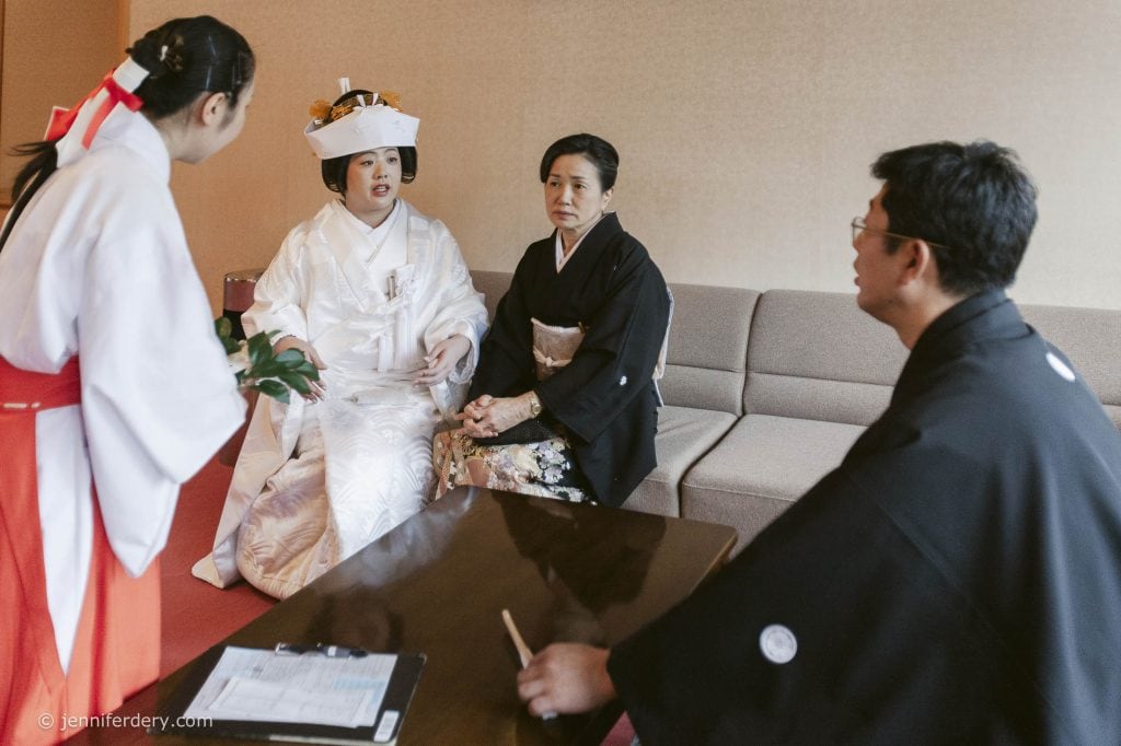 A Japanese bride in a traditional white kimono and headdress sits with her mother and another man in black formal attire while a woman in a red and white ceremonial outfit stands before them in a room with a sofa and table.