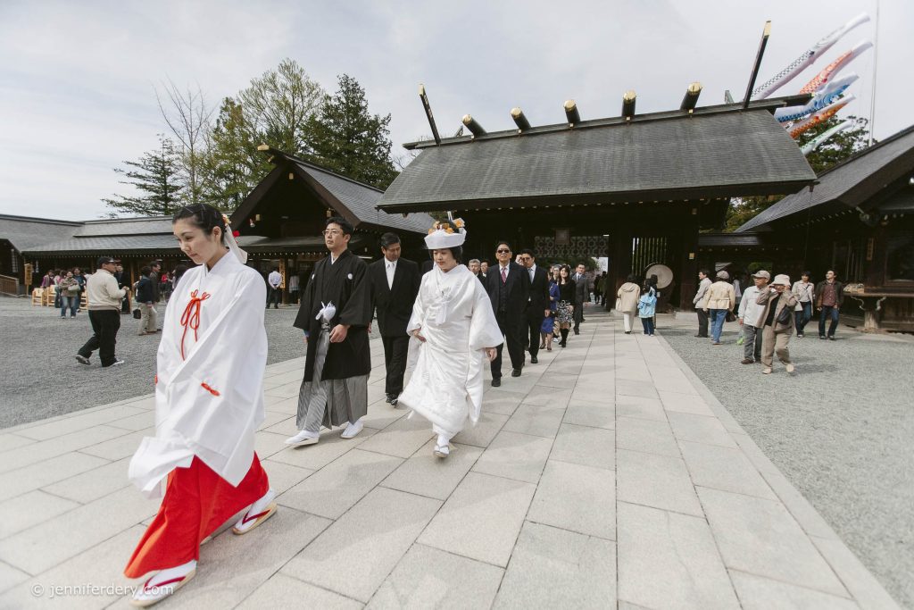 A traditional Japanese wedding procession, with the bride in a white kimono and headdress, groom in black, and attendants in ceremonial attire, walking outside a Shinto shrine as onlookers watch.