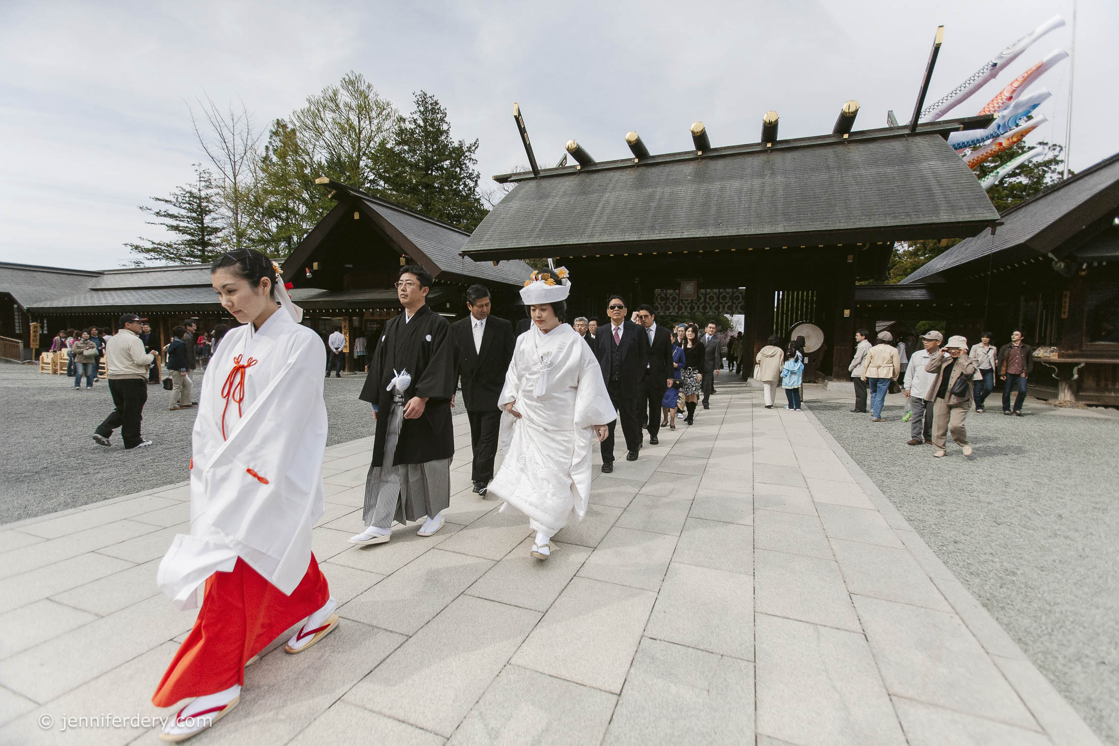 A traditional Japanese wedding procession, with the bride in a white kimono and headdress, groom in black, and attendants in ceremonial attire, walking outside a Shinto shrine as onlookers watch.