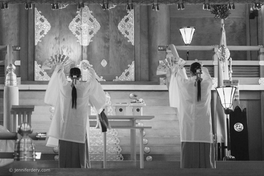 Two people in traditional Japanese attire perform a ritual dance with fans inside a wooden shrine. Ornate decorations and lanterns are visible in the background. The image is in black and white.