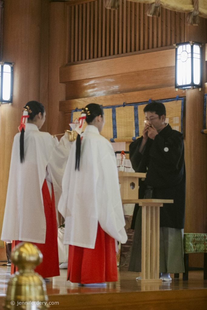 Two women in traditional white and red attire serve a drink to a man in black robes inside a Japanese shrine, with wooden walls and soft natural lighting.