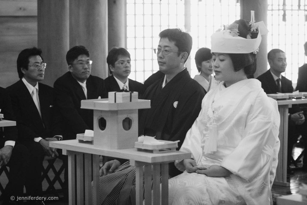 A bride and groom in traditional Japanese wedding attire sit side by side during a ceremony, with guests dressed formally and wooden tables in front of them. The setting appears solemn and respectful.