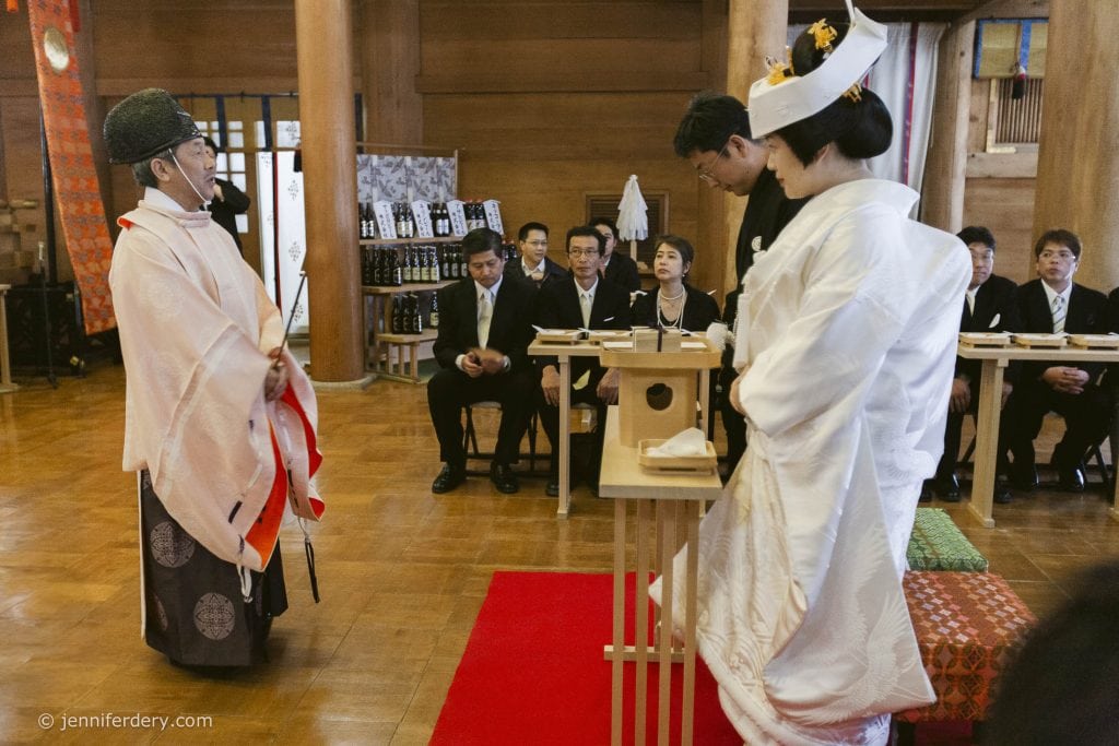 A bride and groom in traditional Japanese wedding attire stand before an officiant in a wooden hall, with seated guests in formal black suits observing the ceremony.