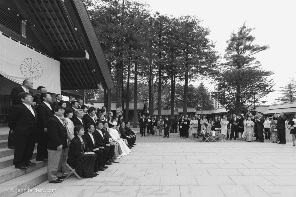 A large group of people, many in traditional Japanese attire, pose for a photo on temple steps while others gather nearby outdoors, surrounded by tall trees and modern buildings in the background.