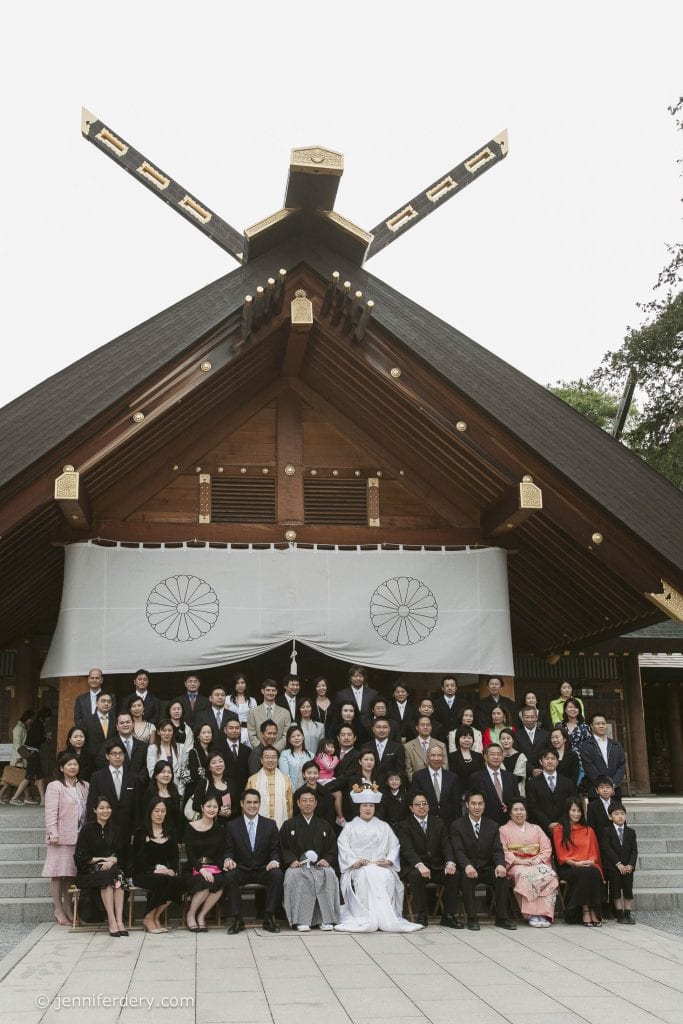 A large group of people, dressed formally, pose for a photo on the steps of a traditional Japanese shrine. The bride and groom sit in the front row, surrounded by family and friends. The shrine features wooden architecture and a white curtain.