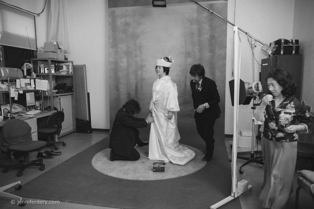 A bride in traditional Japanese attire stands in a photography studio while three people assist with her dress and appearance. Studio equipment and shelves with items are visible in the background. The scene is in black and white.