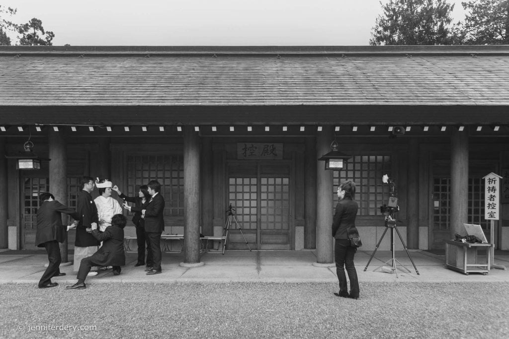 A group of people prepares a person dressed in traditional Japanese attire in front of a wooden building, while another person stands nearby with camera equipment. The scene appears to be a photoshoot. The image is in black and white.