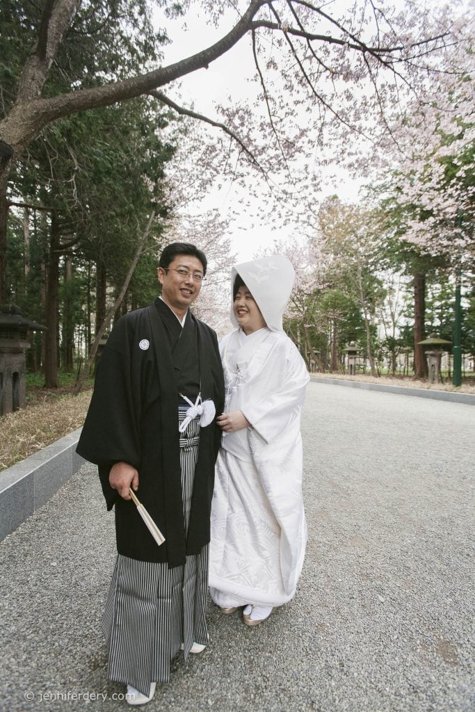 A couple in traditional Japanese wedding attire stands outdoors under cherry blossom trees, smiling at each other. The groom wears a black kimono, and the bride wears a white kimono with a large headpiece.