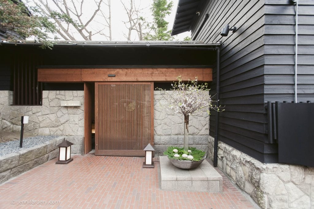 A modern Japanese entrance features a wooden slatted door, stone walls, brick pathway, two lanterns, and a large planter with flowering branches and greenery. The building exterior is black with wood and stone accents.