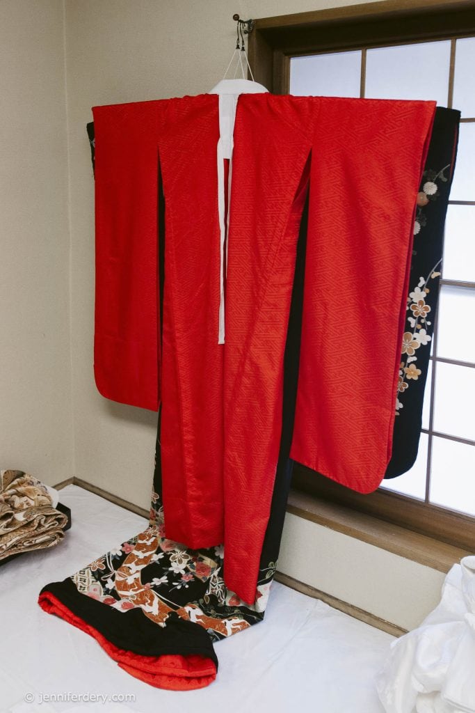A vibrant red and black Japanese kimono with floral patterns hangs on a hanger in a traditional room, next to a window with wooden lattice, draping elegantly onto a white surface.
