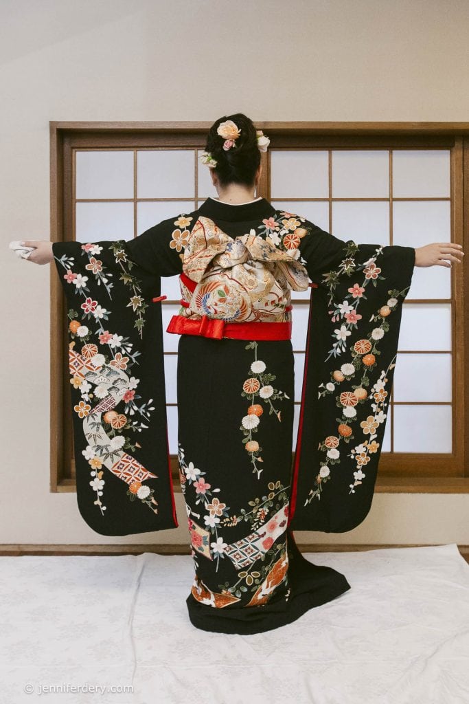 A person stands with arms outstretched, wearing a black kimono with colorful floral and geometric patterns and a decorative obi sash, in front of a traditional Japanese shoji window.