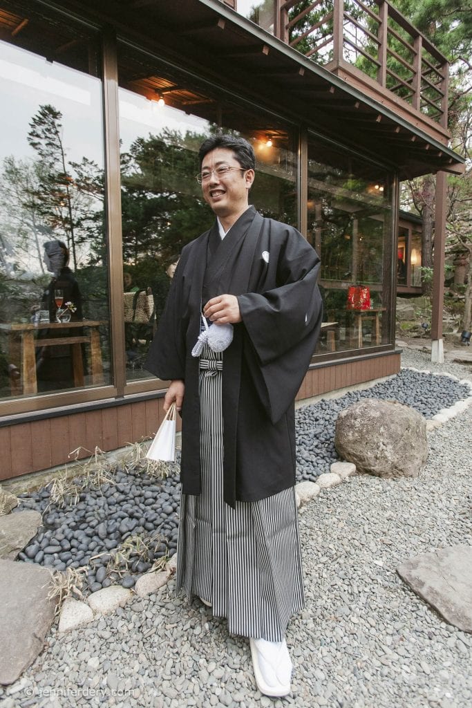 A man wearing traditional Japanese formal attire, including a black kimono, hakama with vertical stripes, and white tabi socks, stands outside a wooden building surrounded by rocks and trees, smiling and holding a fan.