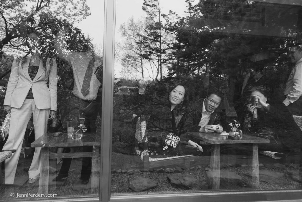 A black-and-white photo shows people indoors seen through a glass window. Three people are sitting at a table, smiling and waving. Reflections of trees and other figures standing outside appear on the glass.