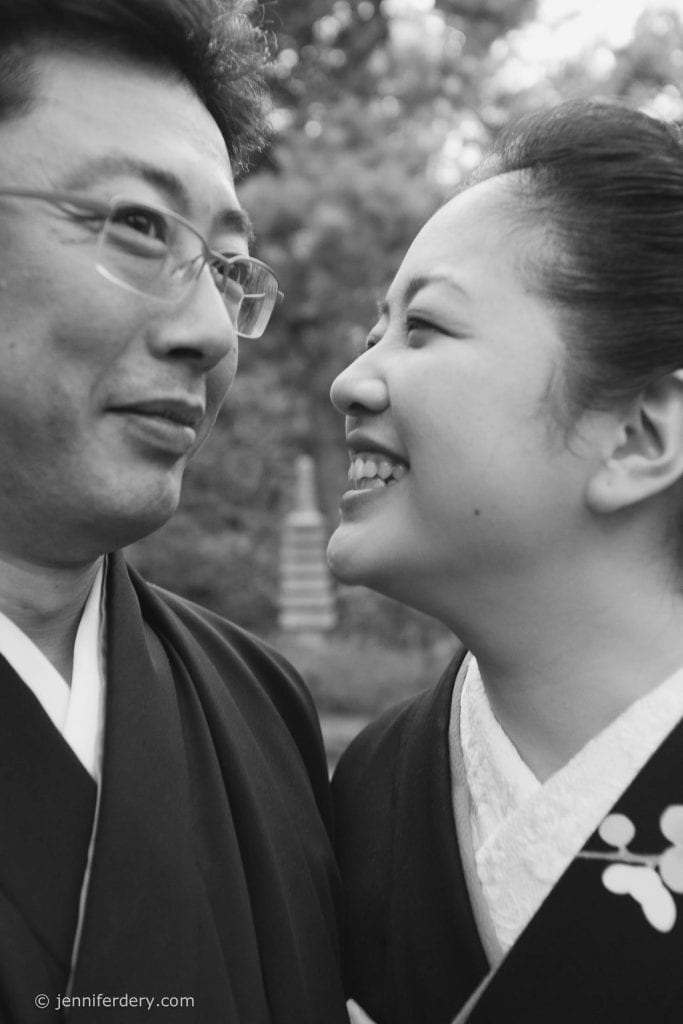 A black and white photo of two people wearing traditional clothing. One person smiles joyfully at the other, who looks back with a gentle expression. The background is slightly blurred, showing outdoor greenery.
