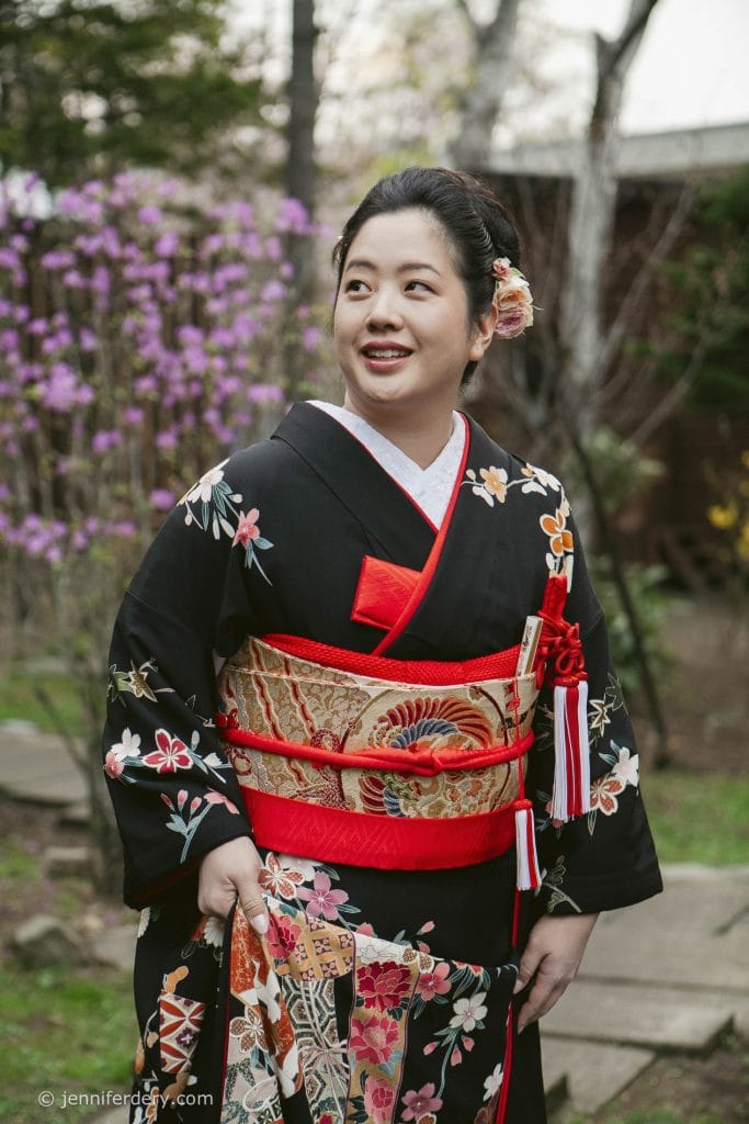 A woman stands outdoors wearing a black kimono with colorful floral patterns and a gold and red obi belt. She has a pink flower in her hair and is smiling, with blooming trees and greenery in the background.