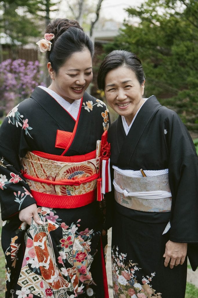 Two women wearing black floral kimonos stand close together outdoors, smiling warmly. One wears a red and gold obi and has a flower hair accessory; the other wears a silver obi. Green trees and purple flowers are in the background.
