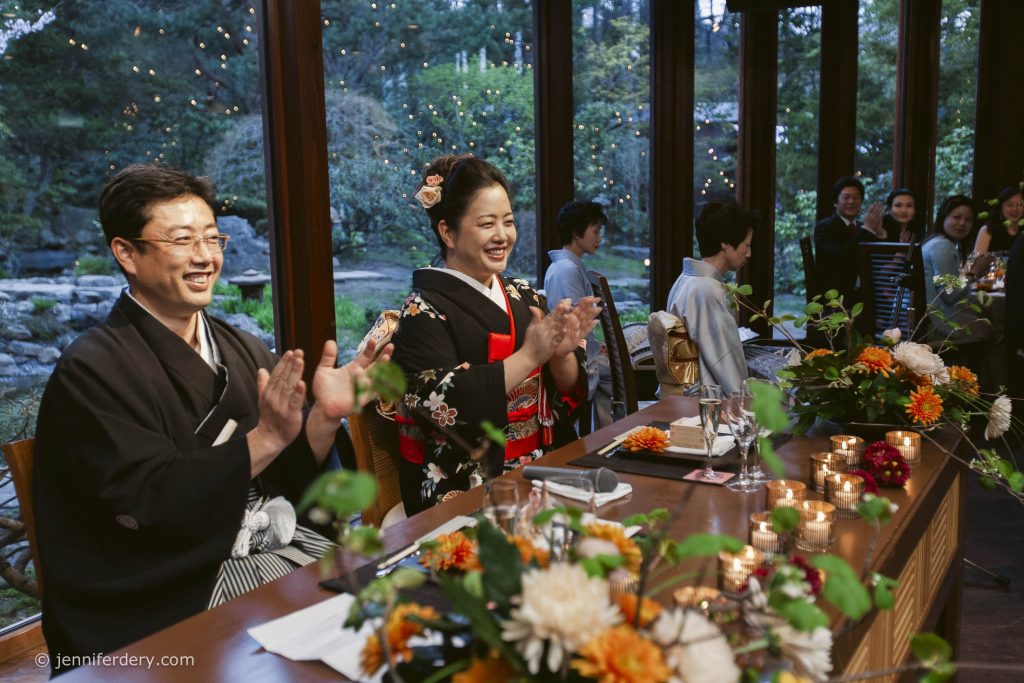 A bride and groom in traditional Japanese attire sit at a decorated table, smiling and clapping. Guests and floral arrangements surround them, with a garden visible through large windows in the background.