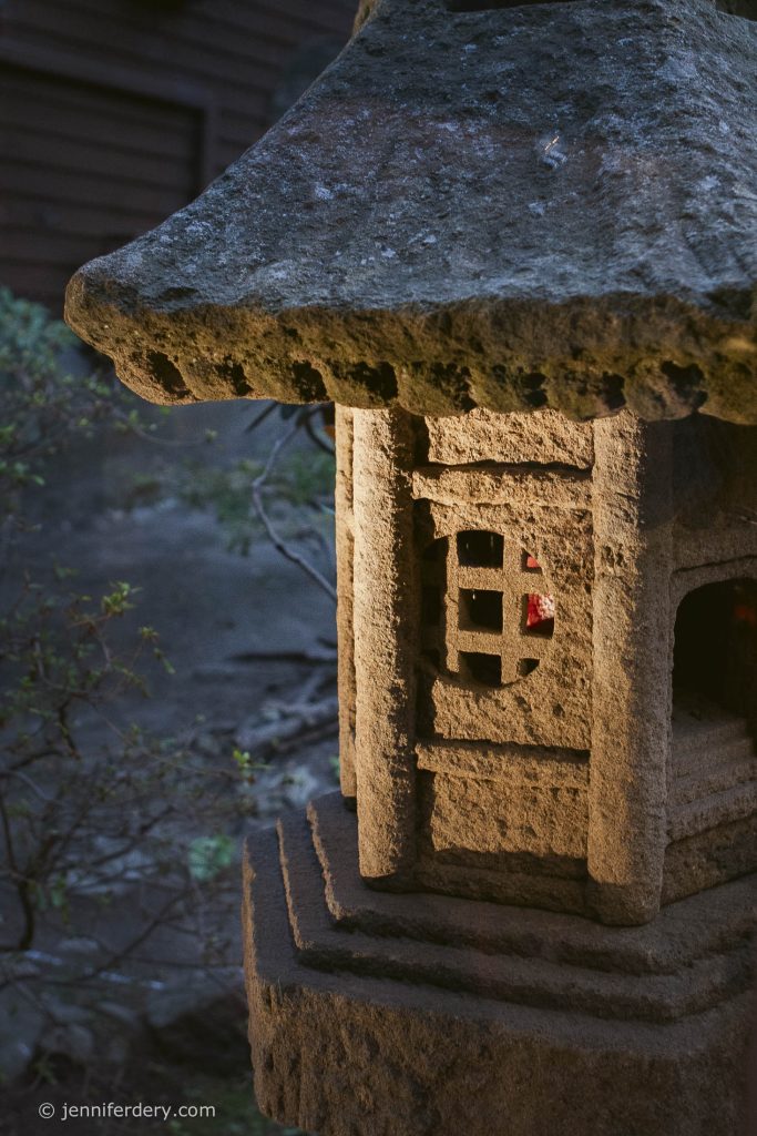 A close-up of a stone lantern with a square window and weathered roof, illuminated by soft light, in a garden setting. The background is out of focus with greenery and dark structures.