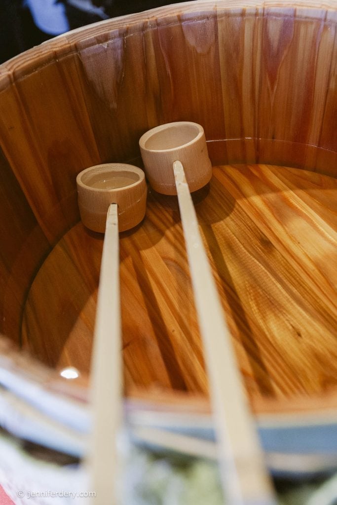 Two wooden ladles rest inside a round wooden basin filled with water. The basin has a smooth, polished finish, highlighting the natural wood grain.
