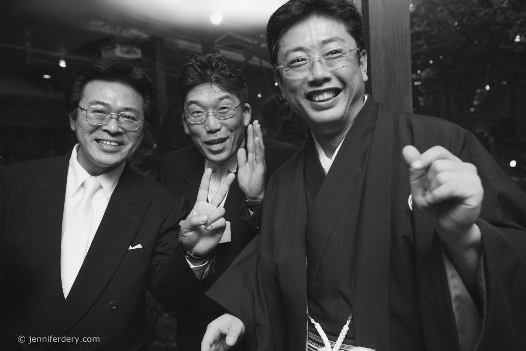 Three men in suits and glasses smile and gesture cheerfully at the camera; one wears traditional Japanese attire. The atmosphere is lively and friendly in this black-and-white photo.