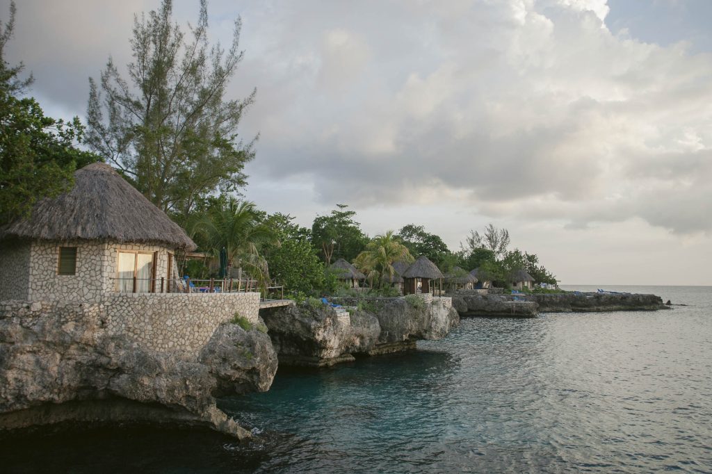 Rockhouse Hotel, Negril, Jamaica: Stone cottages with thatched roofs sit on rocky cliffs above clear blue water, surrounded by lush palm trees and greenery under a partly cloudy sky at sunset.