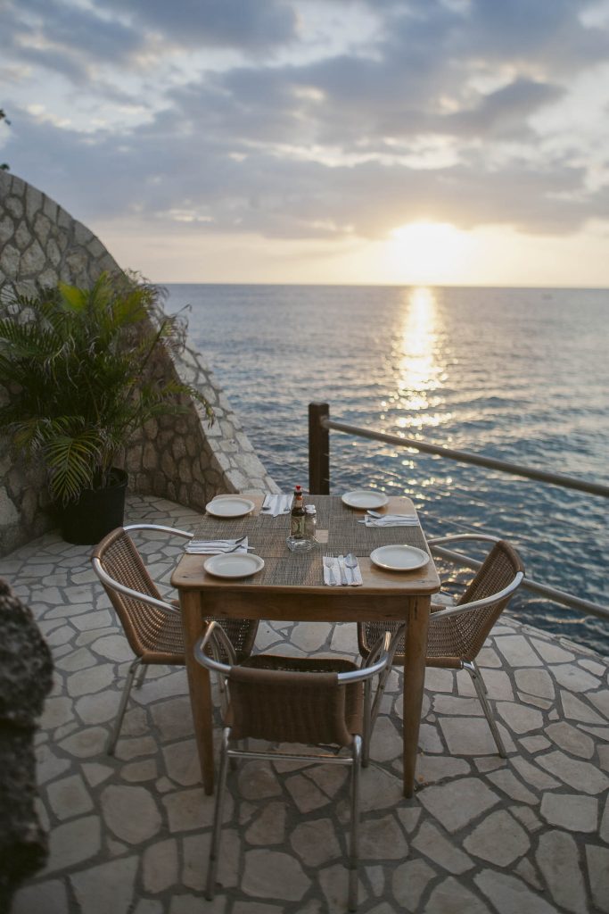 Rockhouse Hotel, Negril, Jamaica: A wooden table set for four with plates and cutlery overlooks the ocean at sunset on a stone patio, creating a serene and romantic outdoor dining setting.