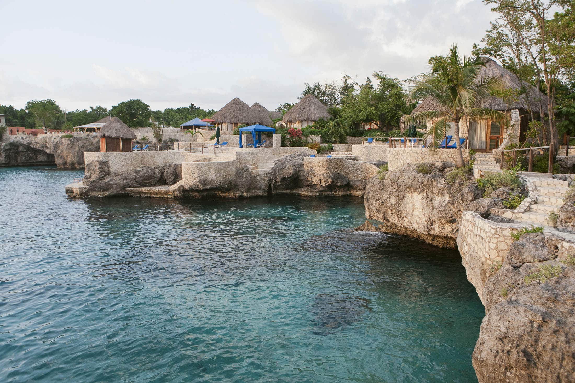 Rockhouse Hotel, Negril, Jamaica: Rocky coastline with clear turquoise water, stone patios, thatched-roof huts, lounge chairs, and palm trees. The area looks peaceful and tropical, with greenery and a few buildings in the background.
